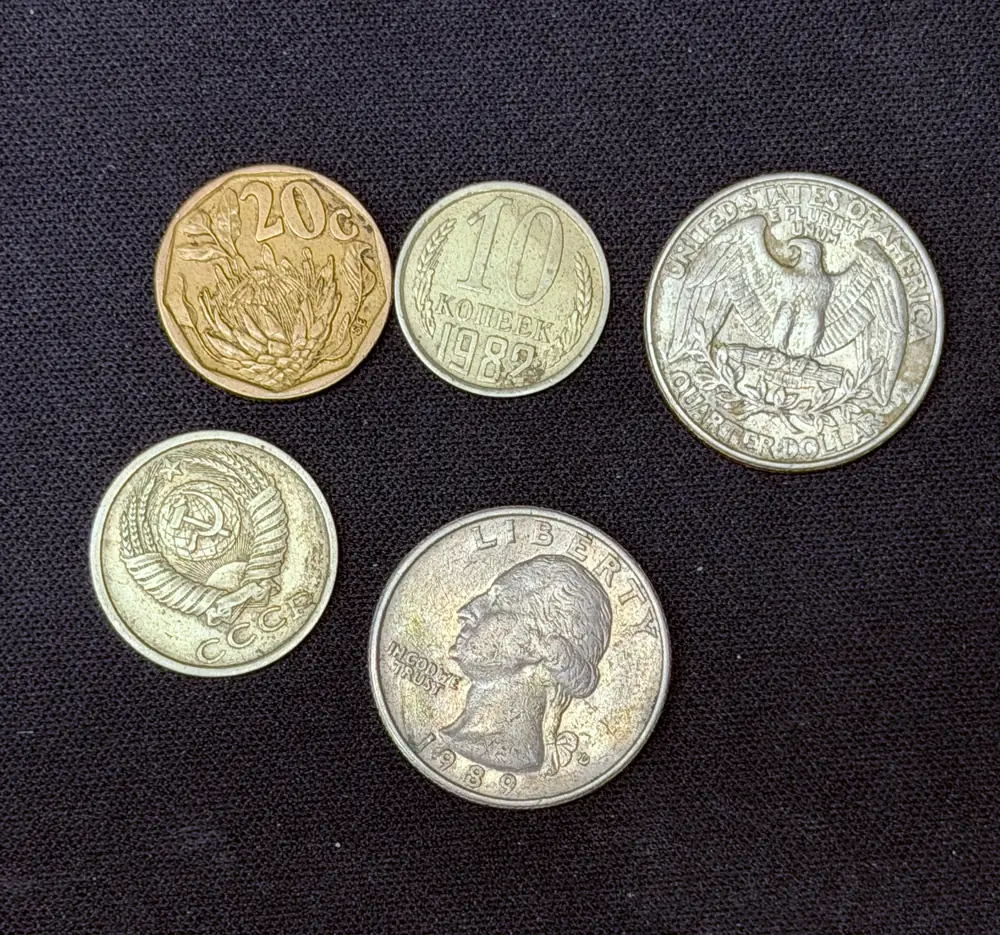 Five worn coins laid out on dark cloth: a brass South African 20 cent, a Soviet 10 kopeck dated 1982, the eagle reverse of a US quarter dollar, a Soviet coin showing the СССР state emblem, and the 1989 obverse of a US quarter. The quarters are visibly wider than the kopecks.