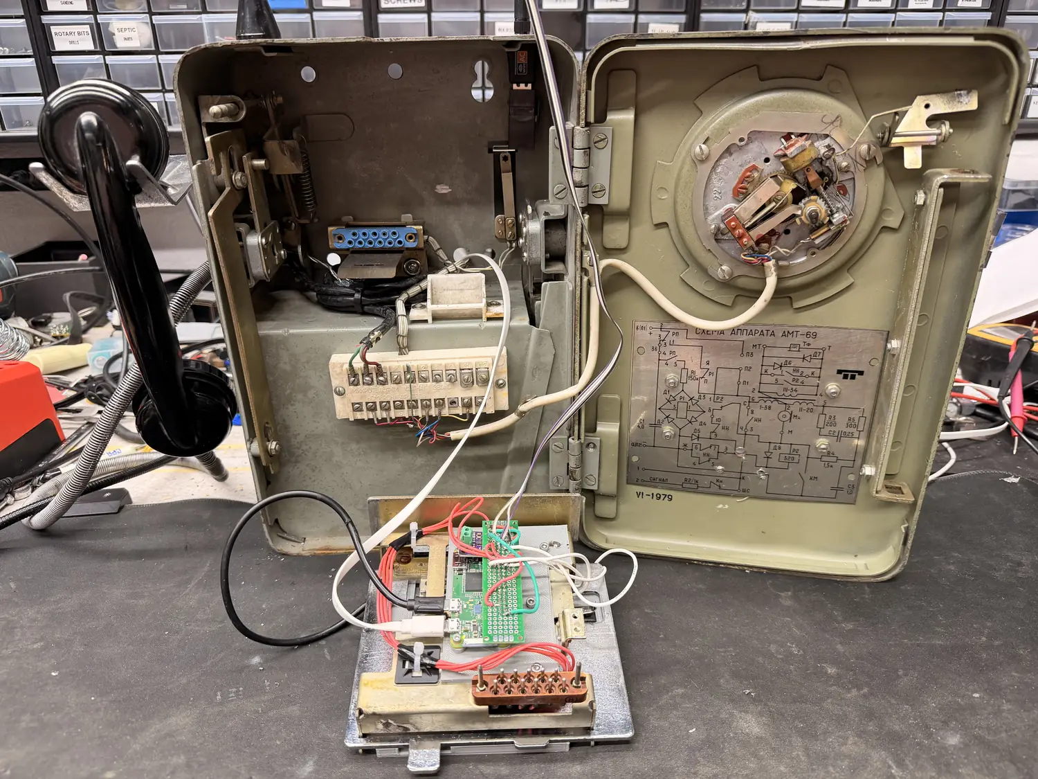 The payphone open on a workbench, both halves laid flat. The left half holds the porcelain terminal block and the original wiring; the right half carries the rotary dial mechanism and the AMT-69 schematic plate stamped 1979. Below them sits the printed plate with the Pi Zero, wired up into both halves.
